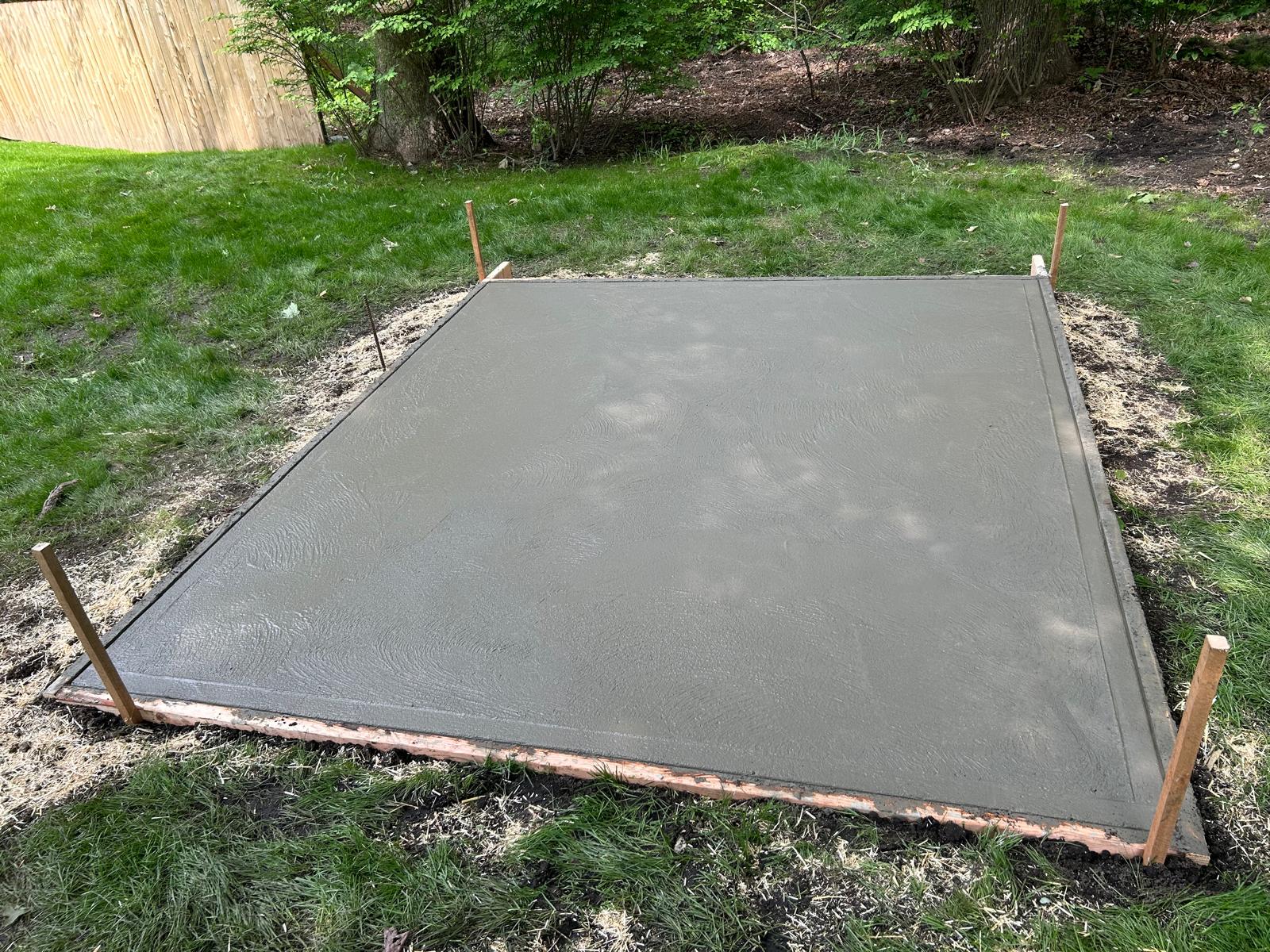 A freshly poured square concrete slab with a broom finish in a grassy backyard