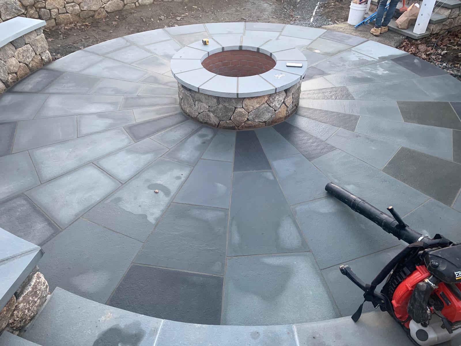 Circular outdoor stone fire pit surrounded by blue and gray tiled seating area with a leaf blower nearby